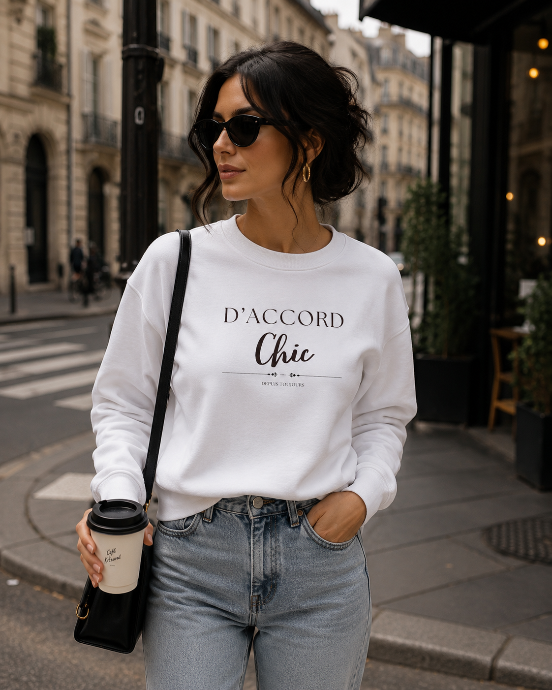 Woman wearing a white D’Accord Chic crewneck sweatshirt with dark front typography, paired with light-wash jeans while standing on a city street.