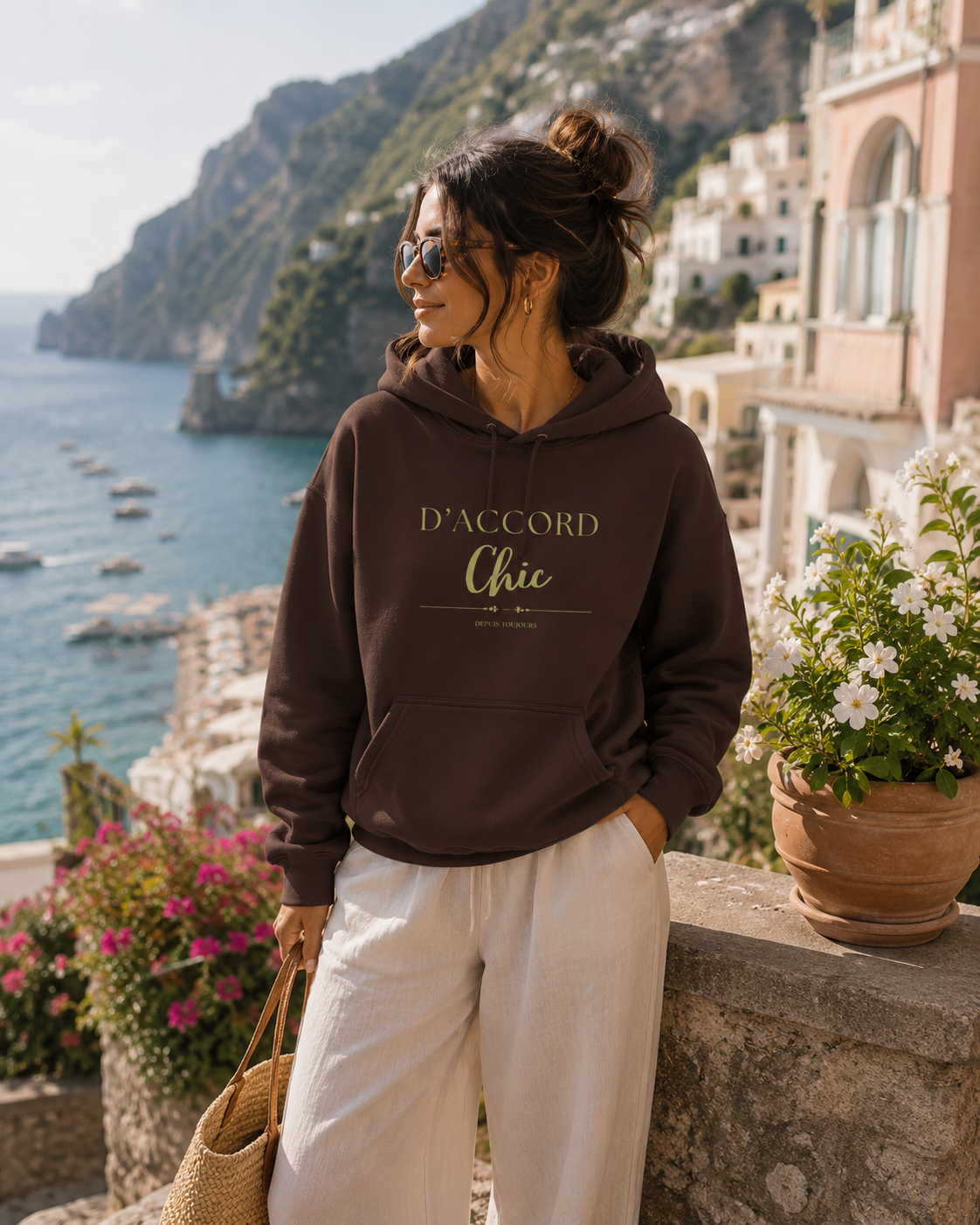 Woman wearing a dark brown D’Accord Chic hoodie with gold front typography, standing on a stone balcony overlooking a coastal town, the blue sea and distant mountains.