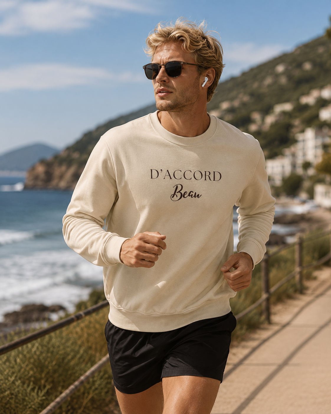 Man jogging on a coastal path wearing a cream D’Accord Beau crewneck sweatshirt with dark chest typography, black shorts and sunglasses, overlooking the blue sea and green hills.
