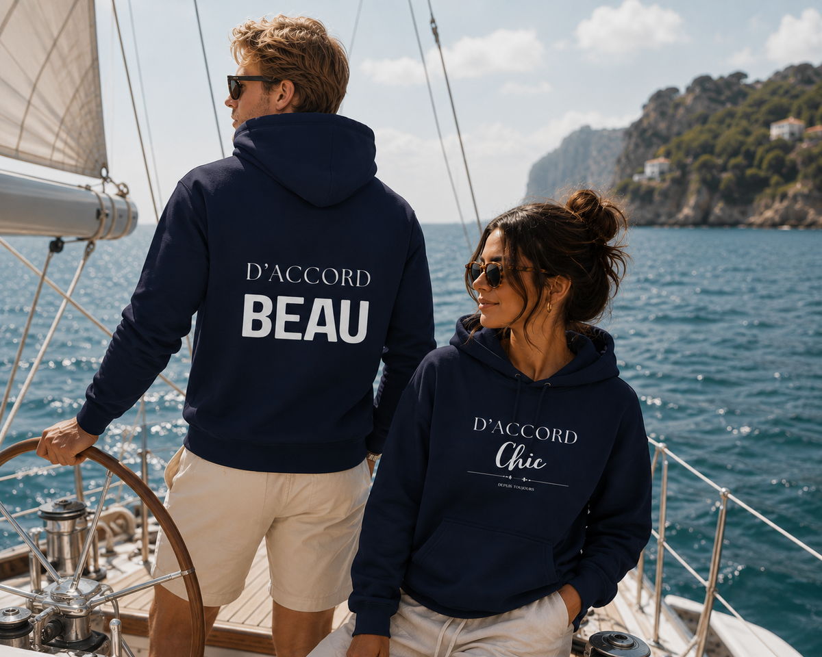 Two models on a sailboat wearing matching navy hoodies. The man wears a D’Accord Beau hoodie with white back typography, while the woman wears a D’Accord Chic hoodie with white front typography.