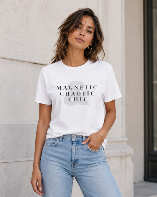 A woman wearing a white graphic t-shirt with the text 'MAGNETIC CHAOTIC CHIC' in black over a light gray circle, paired with light-wash blue jeans, standing against a light beige stone wall.