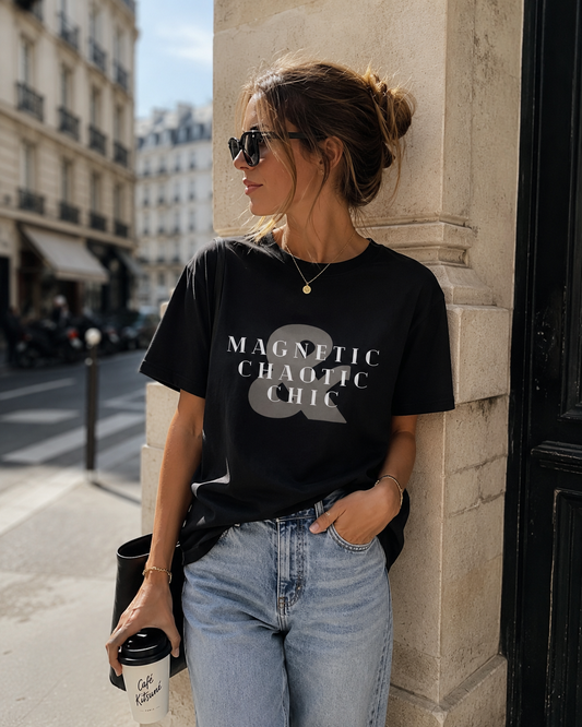 A woman wearing a black graphic t-shirt with the text 'MAGNETIC CHAOTIC CHIC' in white and gray, paired with light-wash blue jeans, leaning against a stone pillar on a city street.
