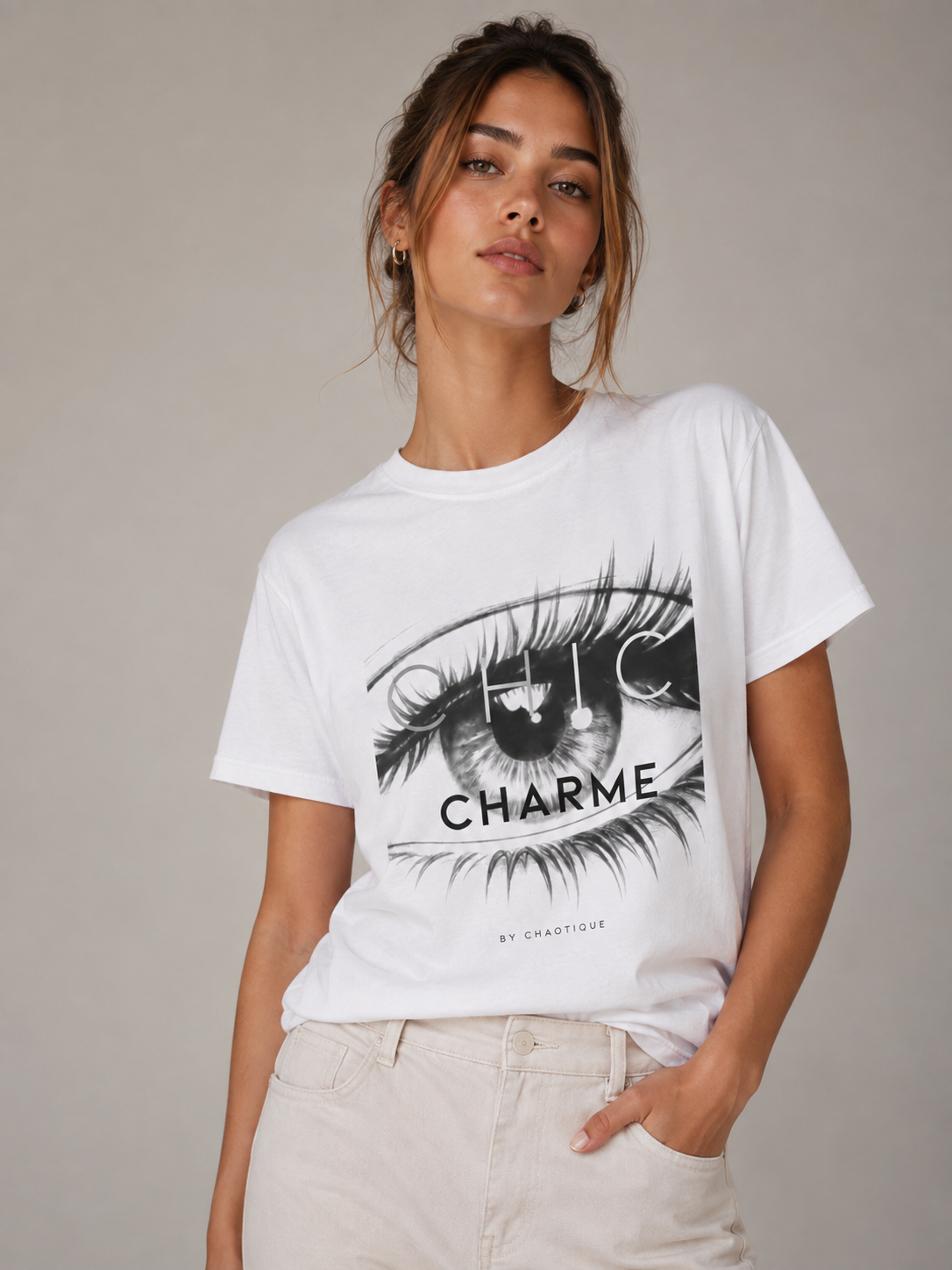 Woman wearing a white CHIC CHARME graphic T-shirt featuring a bold black-and-white eye print, styled with neutral trousers in a minimalist studio setting.