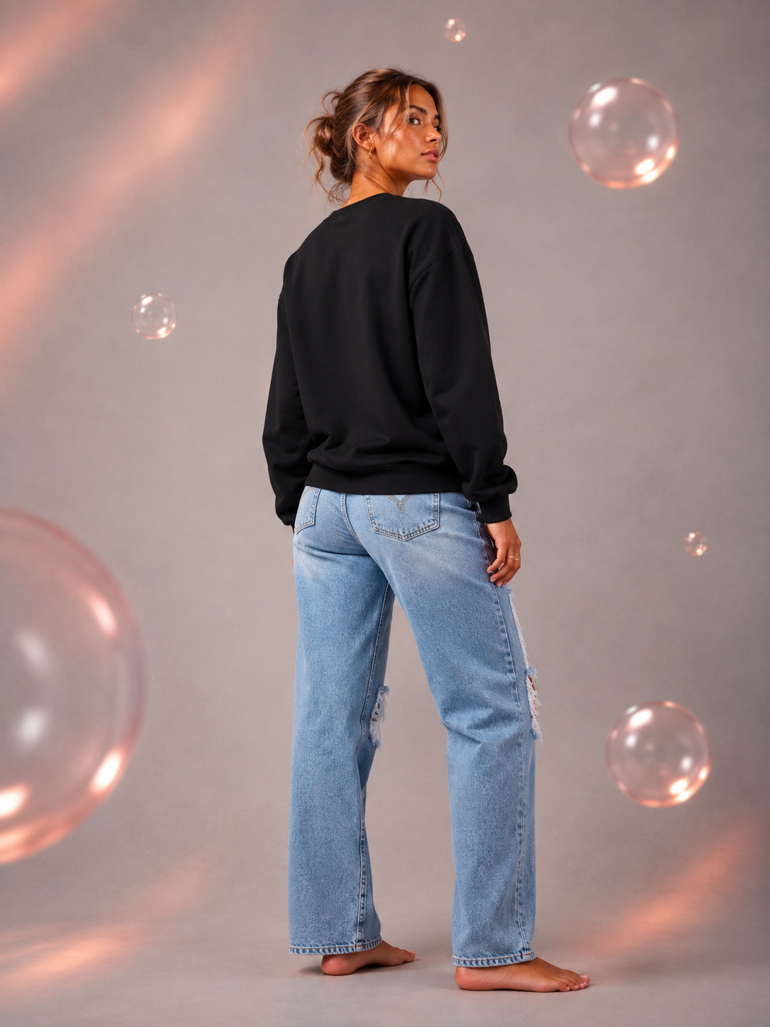 Back view of a woman wearing a black crewneck sweatshirt with light-wash ripped jeans in a gray studio with warm pink lighting and translucent bubbles.