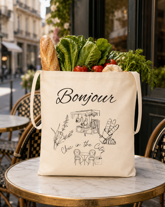 An off-white canvas tote bag with black printed cafe-themed illustrations and the word 'Bonjour' sits on a round cafe table, filled with fresh vegetables and bread, with a blurred outdoor cafe background.
