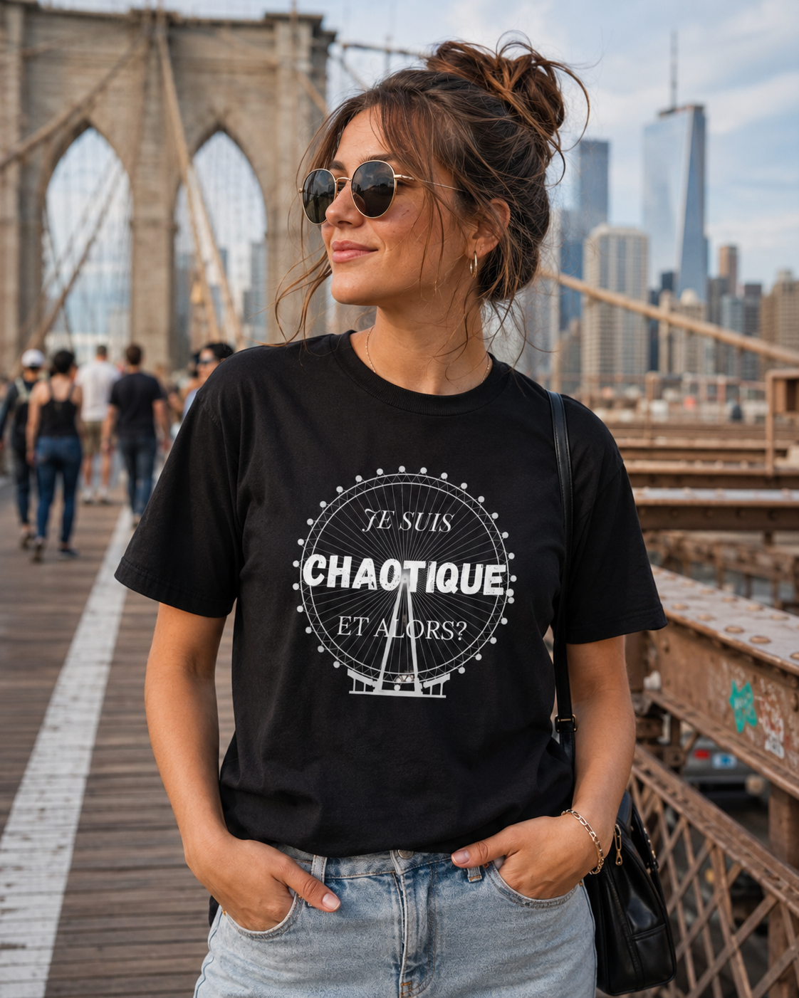 A woman wearing a black graphic t-shirt with a white French text print 'JE SUIS CHAOTIQUE ET ALORS?' in front of the Brooklyn Bridge and New York City skyline.