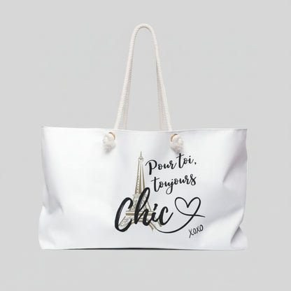 A white canvas tote bag with cream rope handles, featuring a graphic print of the Eiffel Tower and the French text 'Pour toi, toujours Chic XOXO' in black and gold.