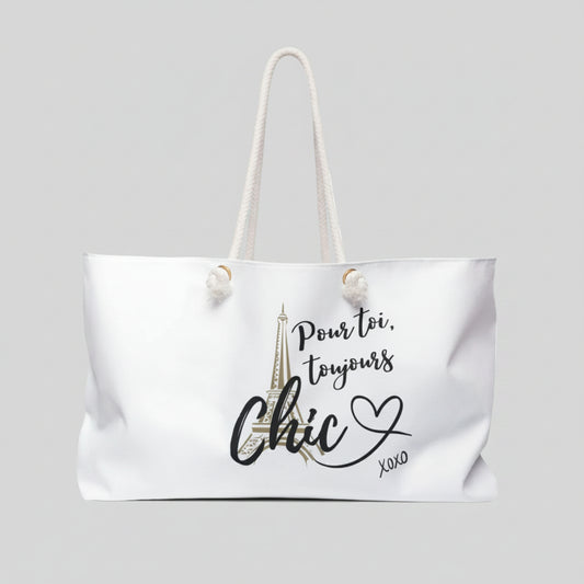 A white canvas tote bag with cream rope handles, featuring a graphic print of the Eiffel Tower and the French text 'Pour toi, toujours Chic XOXO' in black and gold.