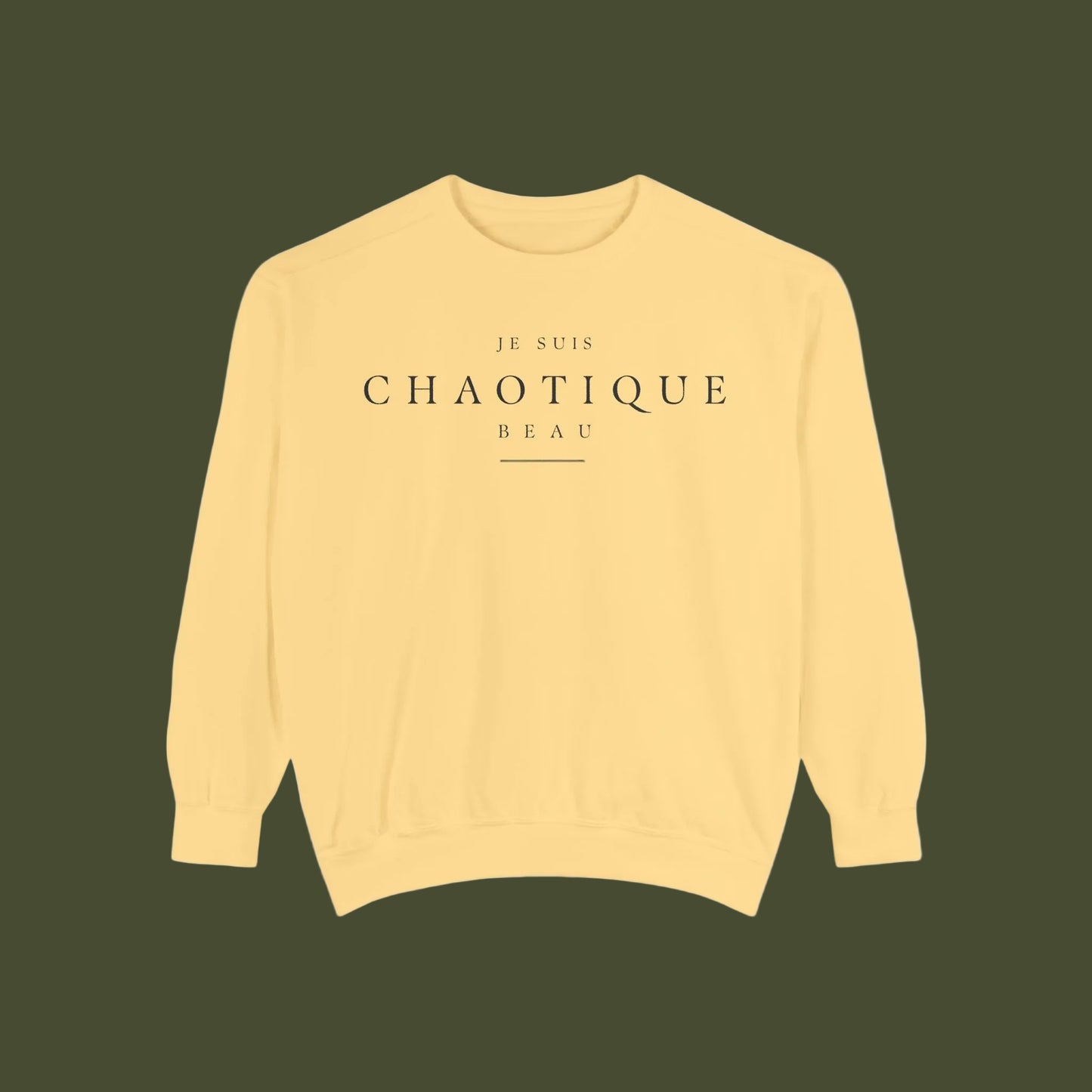 A pale yellow crewneck sweatshirt with 'JE SUIS CHAOTIQUE BEAU' printed in black serif text on the chest, displayed flat against a solid white background.