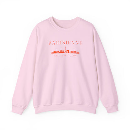 A light pink crewneck sweatshirt with a bright orange “PARISIENNE” graphic, Paris skyline design, and “CHIC À MA FAÇON” text on the chest, displayed against a clean white background.