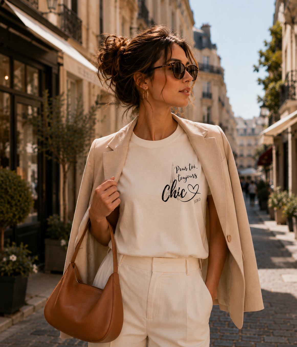 A woman walking down a sunny Parisian street wearing a cream graphic t-shirt with 'Pour Toi toujours Chic' printed on the front, styled with a beige blazer, white trousers, and a brown leather shoulder bag.