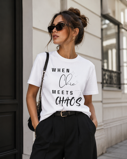 A woman wearing a white graphic t-shirt with the text 'WHEN Chic MEETS CHAOS' in black, styled with black pants, sunglasses, and a crossbody bag, standing outdoors against a light-colored building wall.