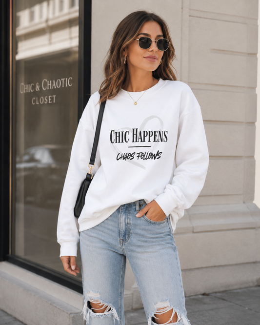 A woman wearing a white crewneck sweatshirt with black graphic text that reads 'CHIC HAPPENS CHAOS FOLLOWS' and light-wash ripped jeans, standing outdoors in front of a building storefront.