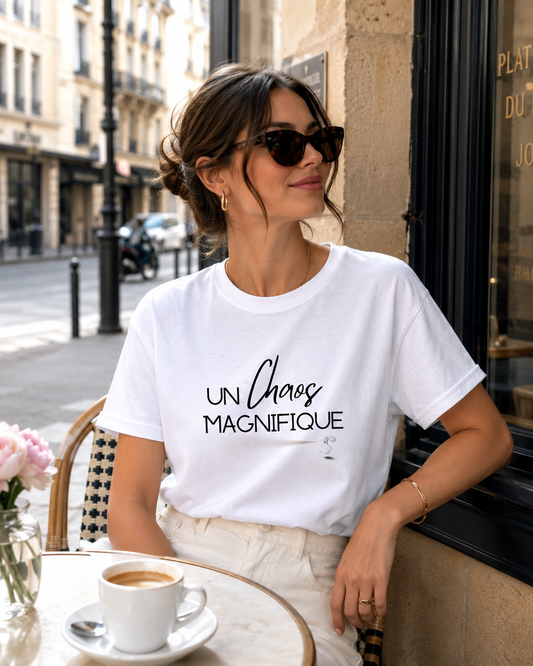 A woman wearing a white graphic t-shirt with the French text 'UN Chaos MAGNIFIQUE' in black script, sitting at an outdoor cafe table with a coffee cup in front of her.