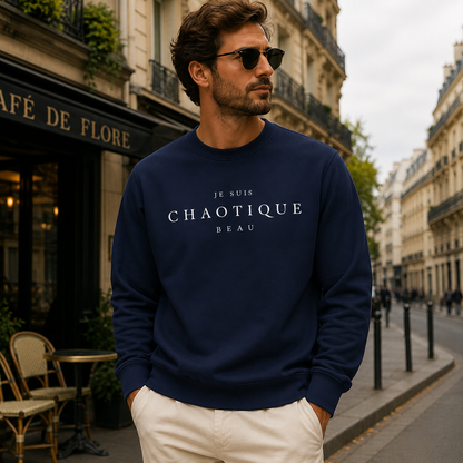A man wearing a navy blue crewneck sweatshirt with 'Je Suis CHAOTIQUE BEAU' printed in white across the chest, paired with off-white trousers, standing on a Parisian street in front of a cafe.