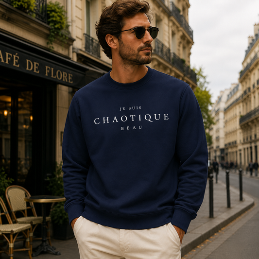 A man wearing a navy blue crewneck sweatshirt with 'Je Suis CHAOTIQUE BEAU' printed in white across the chest, paired with off-white trousers, standing on a Parisian street in front of a cafe.