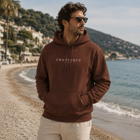 A man wearing a brown hoodie with 'CHAOTIQUE Beau' printed in white on the chest, standing on a beach with the sea and hills in the background.