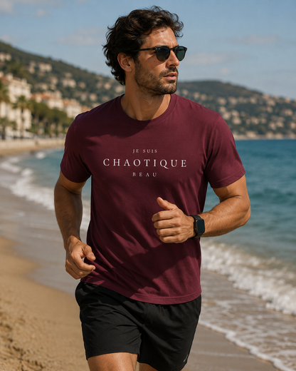 A man wearing a burgundy t-shirt with the text 'JE SUIS CHAOTIQUE BEAU' in white, running on a sandy beach with the blue ocean and hills in the background.