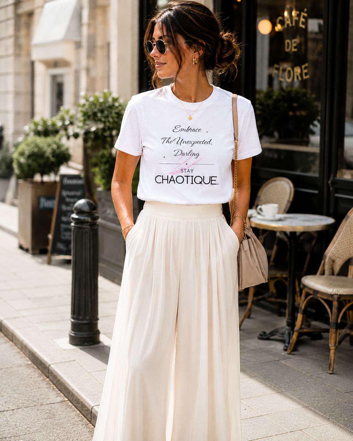 A woman stands on a city sidewalk wearing a white graphic T-shirt with black script text and a long cream pleated skirt. She is wearing sunglasses, a small gold necklace, and a beige shoulder bag, with a cafe storefront in the background.