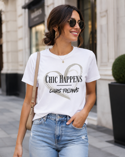 A woman wearing a white graphic t-shirt with a gray heart design and the text 'Chic Happens CHAOS FOLLOWS', paired with light blue jeans and sunglasses, standing on a city street.