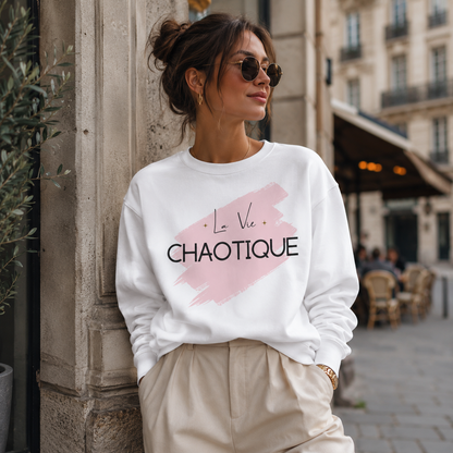 A woman wearing a white crewneck sweatshirt with a pink brushstroke graphic and the text 'La Vie Chaotique' in black, paired with beige trousers, leaning against a stone pillar in an outdoor urban setting.