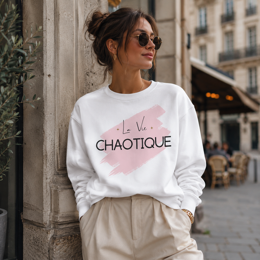 A woman wearing a white crewneck sweatshirt with a pink brushstroke graphic and the text 'La Vie Chaotique' in black, paired with beige trousers, leaning against a stone pillar in an outdoor urban setting.