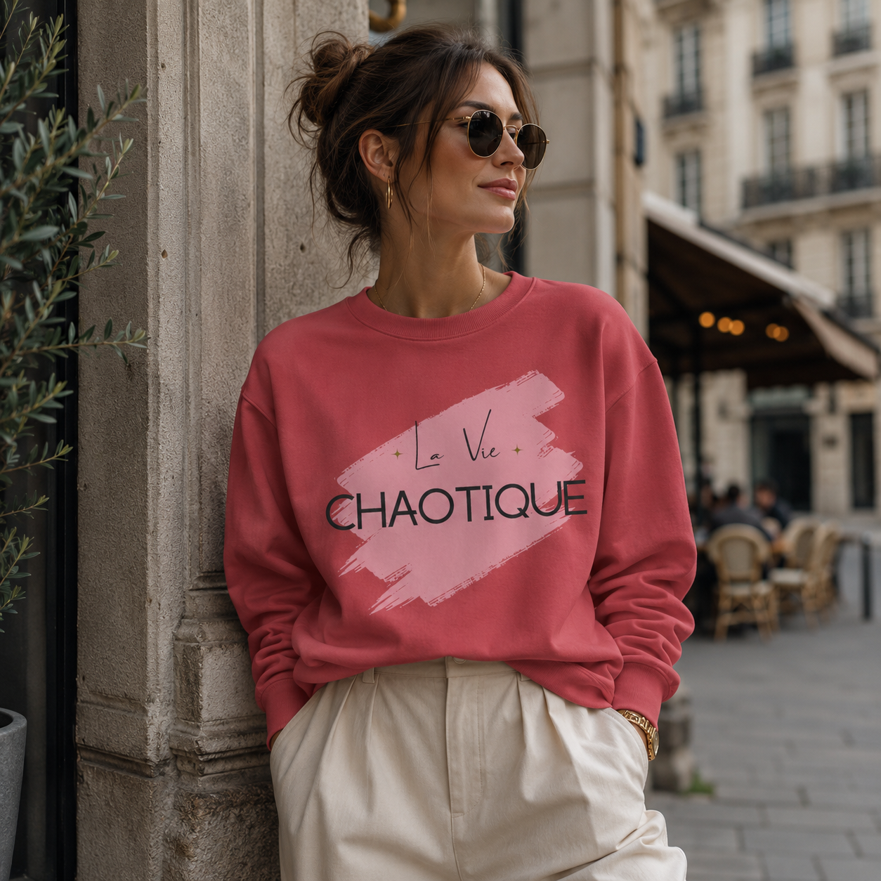 A woman wearing a dusty rose crewneck sweatshirt with a pink brushstroke graphic and the text 'La Vie CHAOTIQUE' in black, paired with cream trousers, leaning against a stone pillar in an outdoor urban setting.