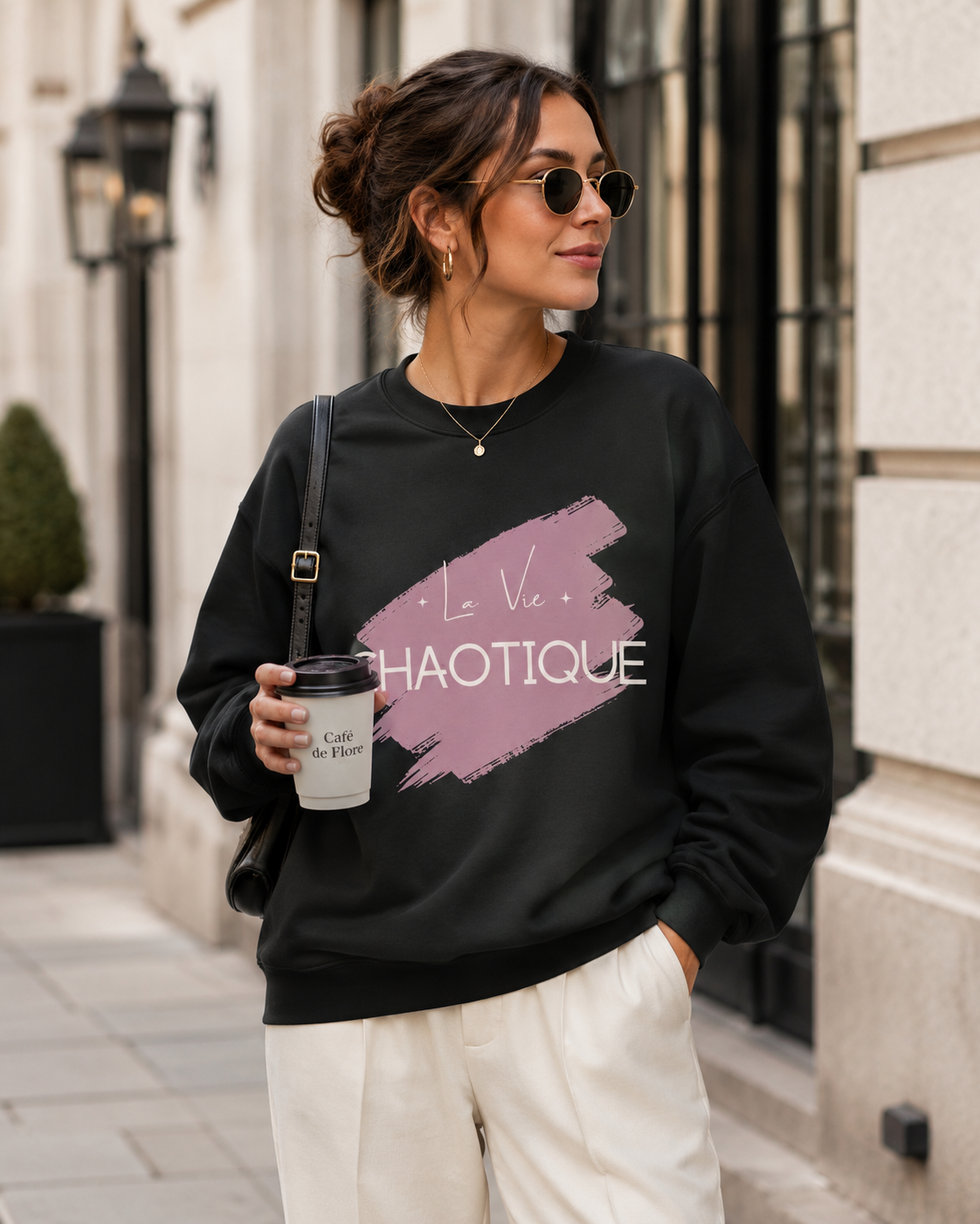 A woman wearing a black crewneck sweatshirt with a pink and white graphic print that reads 'La Vie CHAOTIQUE', styled with cream pants, sunglasses, and a coffee cup in an outdoor urban setting.