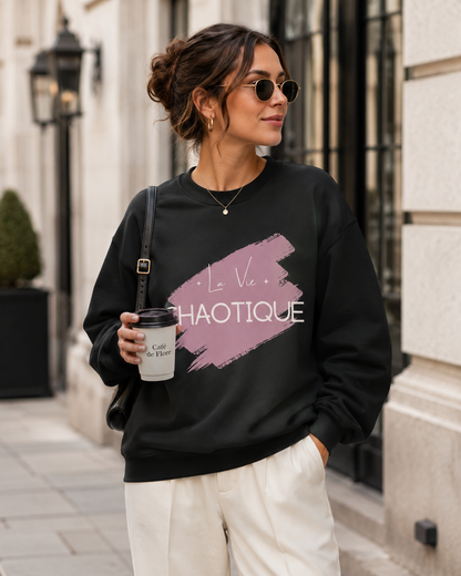 A woman wearing a black crewneck sweatshirt with a pink and white graphic print that reads 'La Vie CHAOTIQUE', styled with cream pants, sunglasses, and a coffee cup in an outdoor urban setting.
