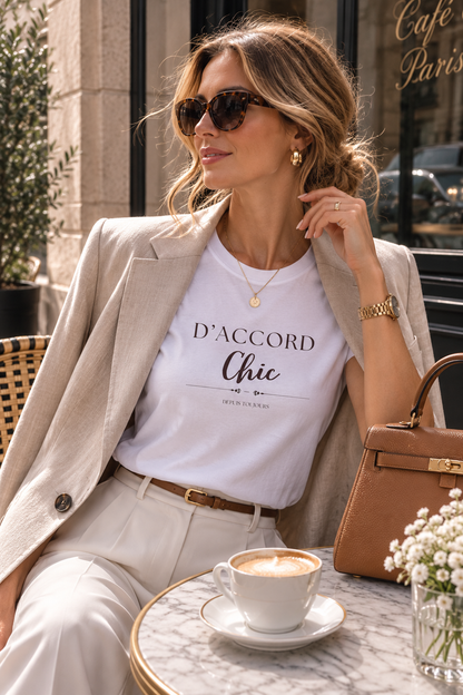 A woman sits at a cafe table wearing a white graphic t-shirt with 'D'ACCORD Chic' printed on the front, styled with a beige blazer, cream trousers, sunglasses, and a brown handbag.