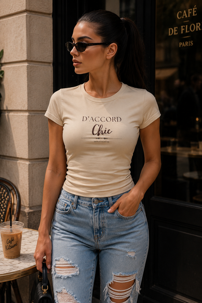 A woman wearing a fitted cream-colored t-shirt with 'D'ACCORD Chic' printed on the front, paired with distressed light-wash blue jeans, standing on a city street in front of a cafe.