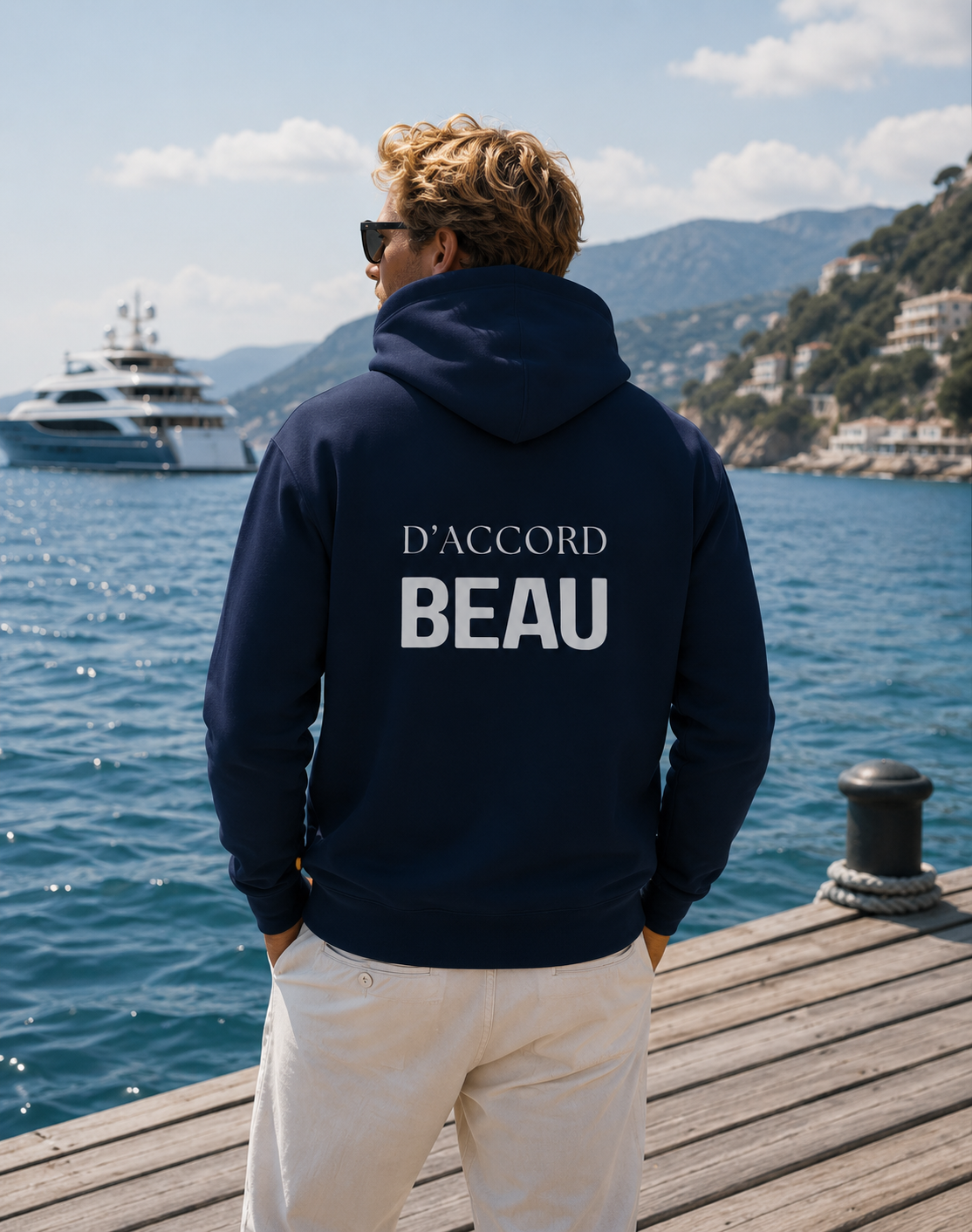 A man stands on a wooden dock overlooking a blue sea and mountains, wearing a navy hoodie from the back with the white text 'D'ACCORD BEAU'.