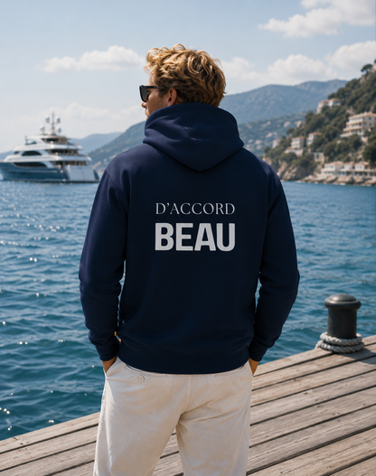 A man stands on a wooden dock overlooking a blue sea and mountains, wearing a navy hoodie from the back with the white text 'D'ACCORD BEAU'.