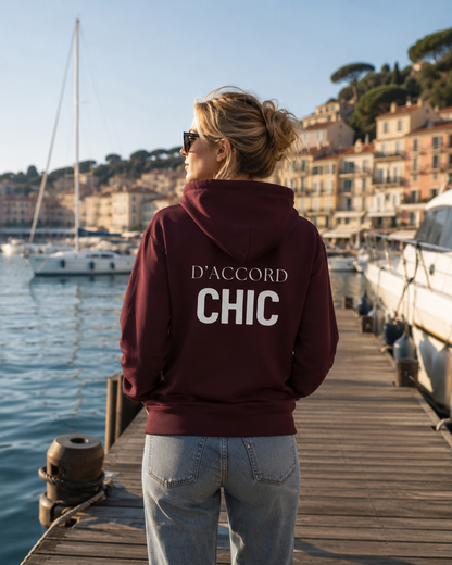 A woman stands on a wooden dock by the water, seen from behind, wearing a maroon hoodie with 'D'ACCORD CHIC' printed in white across the back, paired with light blue jeans. Boats and a coastal town are visible in the background.