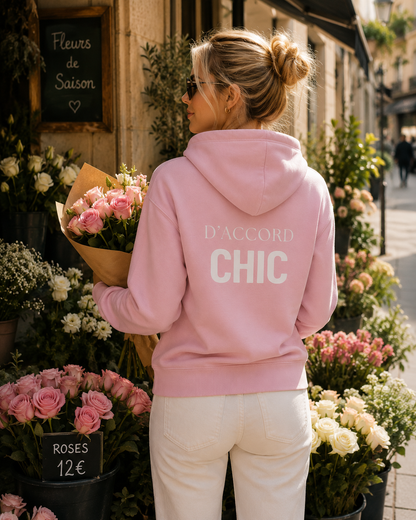 A woman stands with her back to the camera in a flower shop, wearing a light pink hoodie with 'D'ACCORD CHIC' printed in white on the back, paired with white pants.