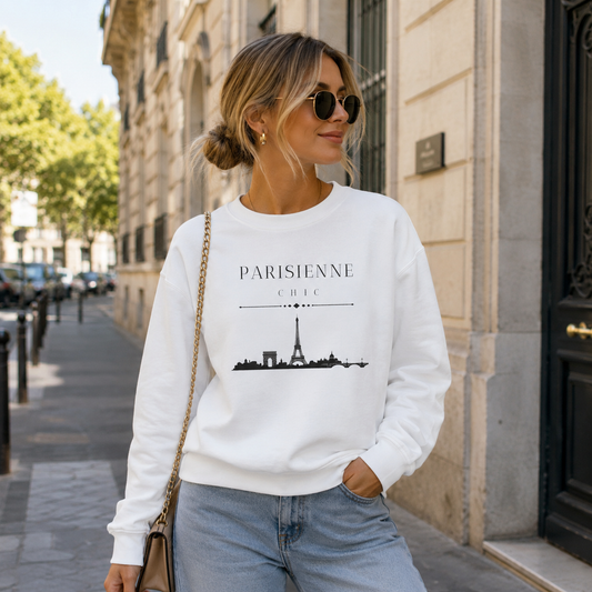 A woman wearing a white crewneck sweatshirt with a black Paris graphic print, paired with light blue jeans and sunglasses, standing on a city sidewalk.