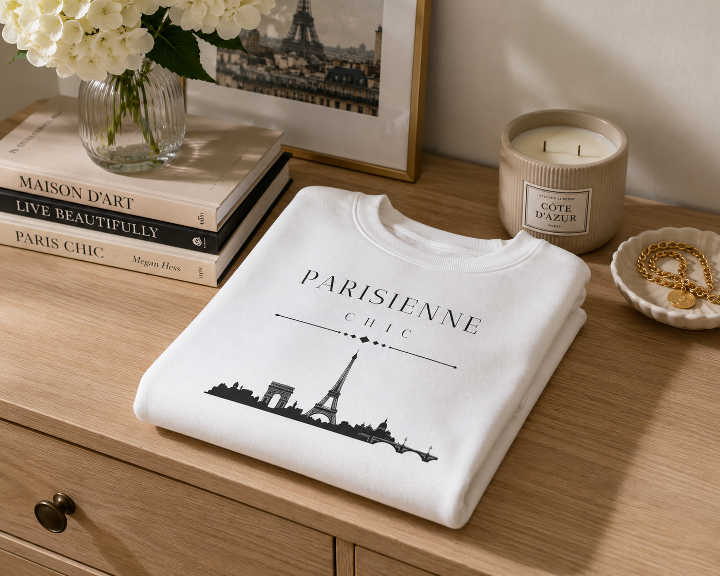A folded white crewneck sweatshirt with a black Paris skyline graphic and the text 'PARISIENNE CHIC' is displayed on a light wood dresser. The scene is styled with books, a vase of white flowers, a framed print, and a candle.