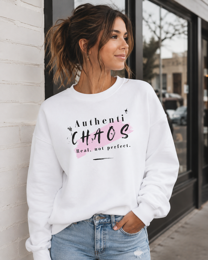 A woman wearing a white crewneck sweatshirt with a pink and black graphic that reads 'AuthentiCHAOS Real, not perfect.' paired with light blue jeans, standing in front of a white brick wall and storefront window.
