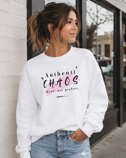 A woman wearing a white crewneck sweatshirt with a pink and black graphic that reads 'AuthentiCHAOS Real, not perfect.' paired with light blue jeans, standing in front of a white brick wall and storefront window.