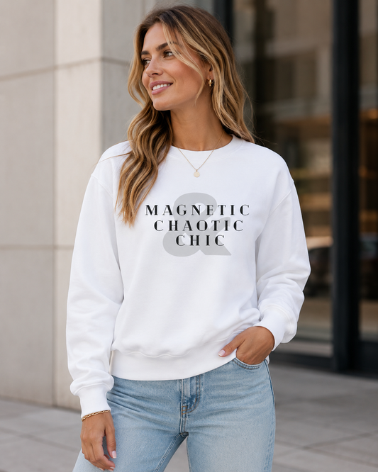 A woman wearing a white crewneck sweatshirt with the text 'MAGNETIC CHAOTIC CHIC' and a large gray ampersand graphic on the front, paired with light blue jeans, standing outdoors in front of a modern building.