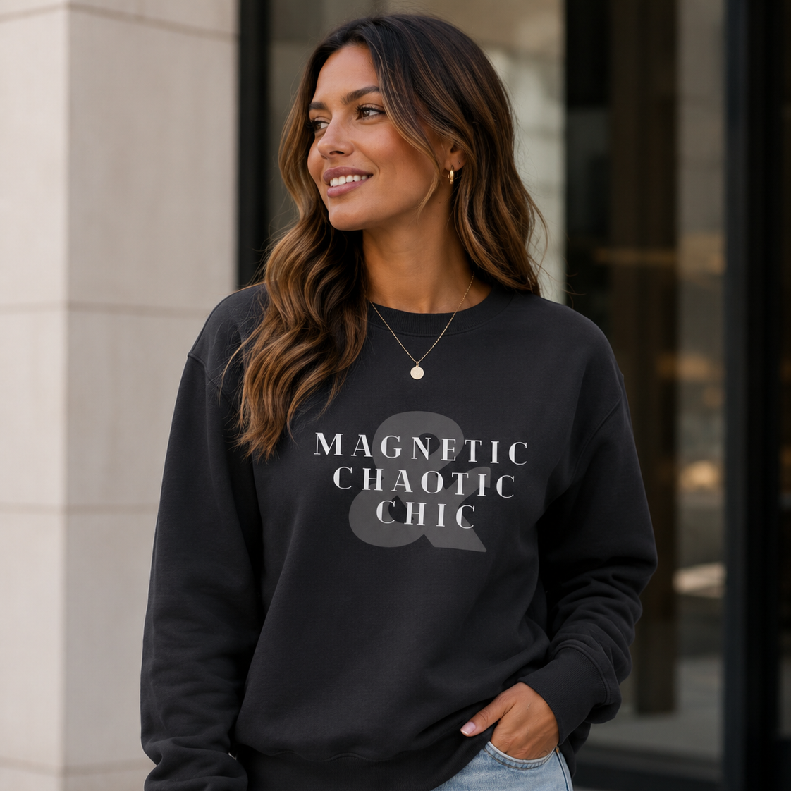 A woman wearing a black crewneck sweatshirt with the text 'MAGNETIC CHAOTIC CHIC' in white, paired with light blue jeans, standing outdoors in front of a modern building.