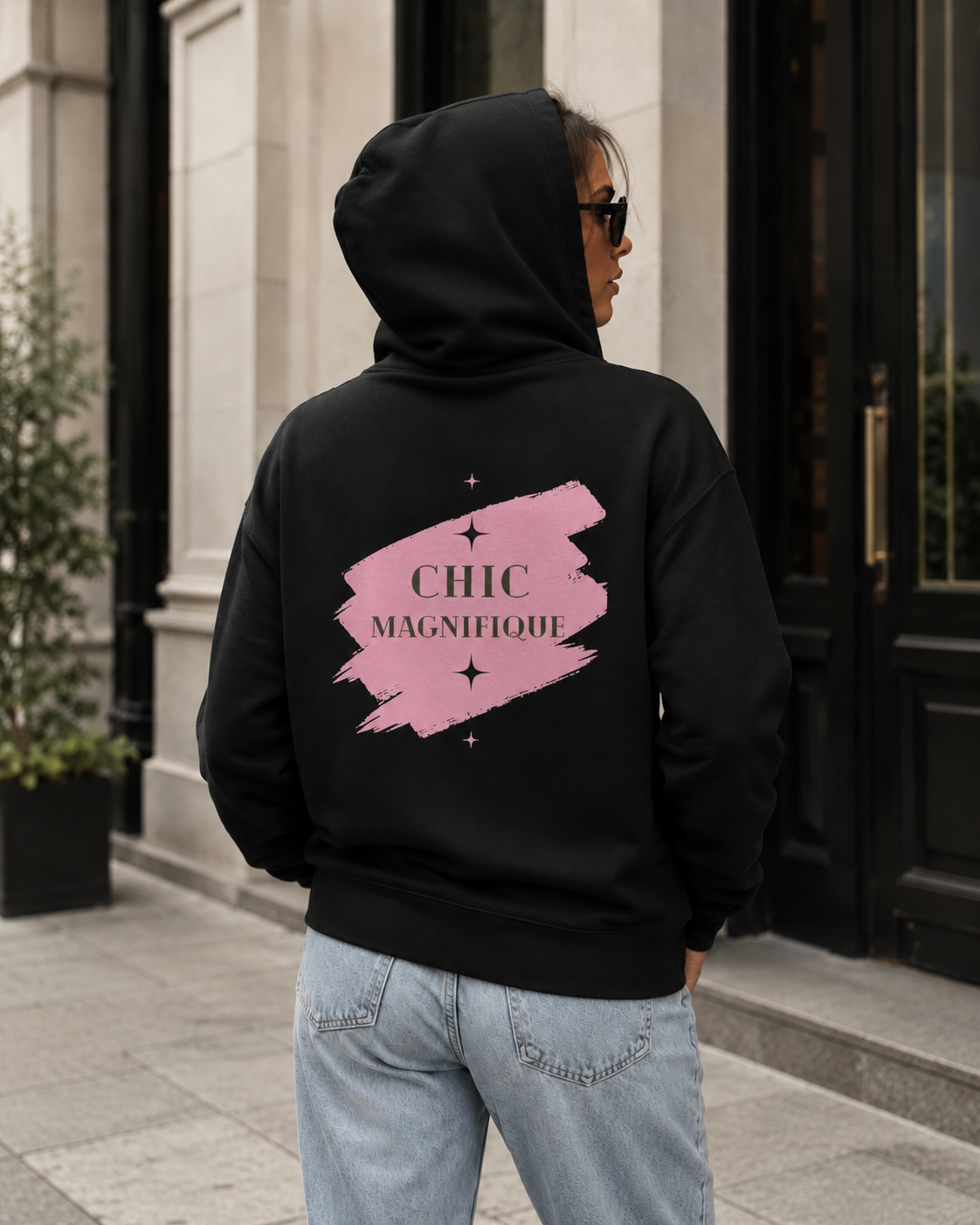 A person wearing a black hoodie with a pink brushstroke graphic and the text 'CHIC MAGNIFIQUE' on the back, standing outdoors in front of a building entrance.