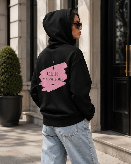 A woman wearing a black hoodie with a pink brushstroke graphic on the back that reads 'CHIC MAGNIFIQUE', paired with light blue jeans, standing outside a building.