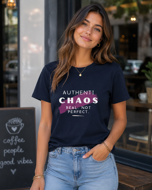 A woman wearing a navy blue graphic t-shirt with the text 'AUTHENTICHAOS REAL, NOT PERFECT.' in white and pink, paired with light wash blue jeans, standing in front of a coffee shop.