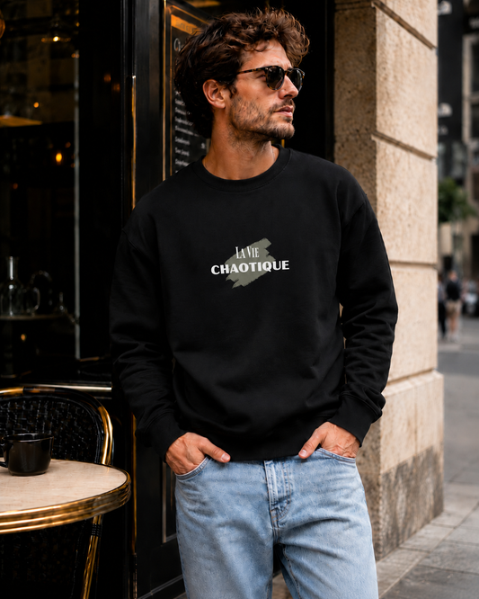 A man wearing a black crewneck sweatshirt with a white graphic print that reads 'La Vie CHAOTIQUE' and light blue jeans, standing outdoors next to a cafe table.