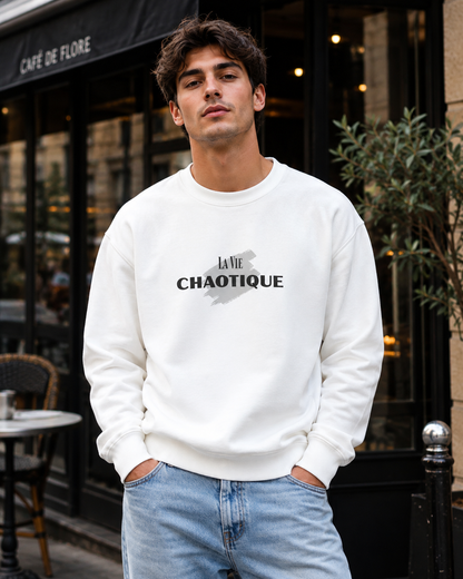 A man wearing a white crewneck sweatshirt with a black graphic print that reads 'LA VIE CHAOTIQUE' and light blue jeans, standing in front of a cafe storefront.