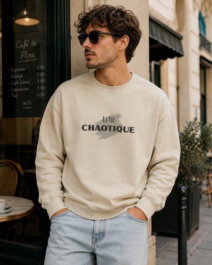 A man wearing a cream-colored crewneck sweatshirt with 'LA VIE CHAOTIQUE' printed in black across the chest, paired with light blue jeans, standing on a city street in front of a cafe.