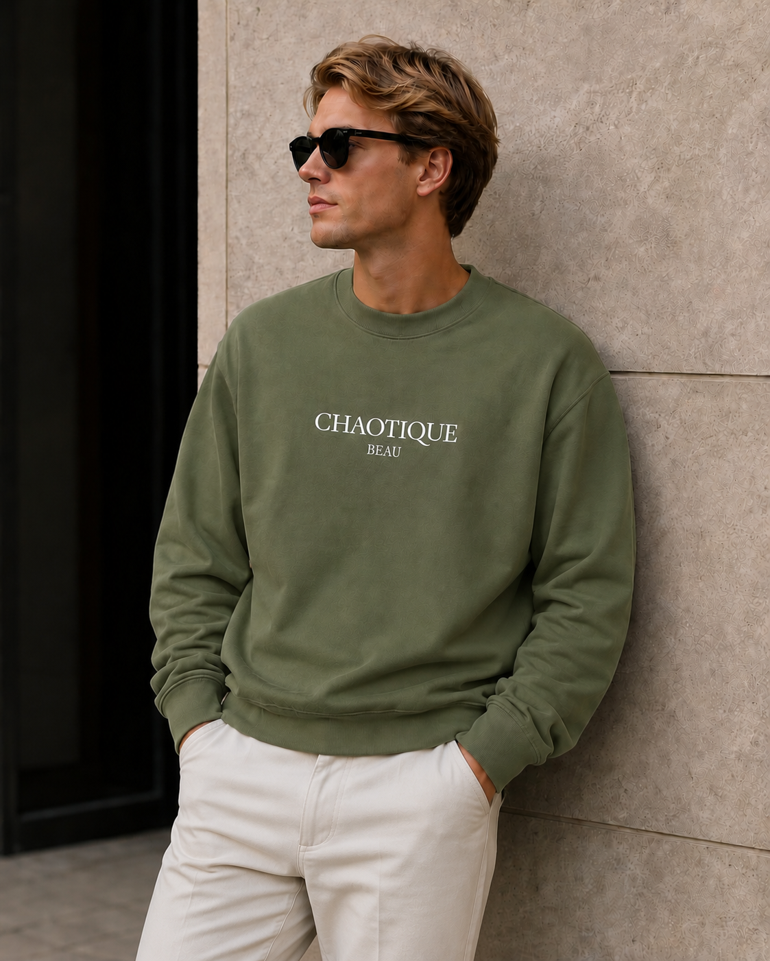 A man wearing an olive green crewneck sweatshirt with 'CHAOTIQUE BEAU' printed in white on the chest, paired with white pants and black sunglasses, leaning against a textured stone wall.