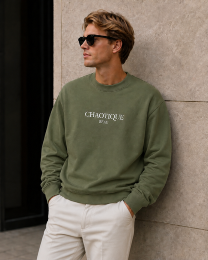 A man wearing an olive green crewneck sweatshirt with 'CHAOTIQUE BEAU' printed in white on the chest, paired with white pants and black sunglasses, leaning against a textured stone wall.