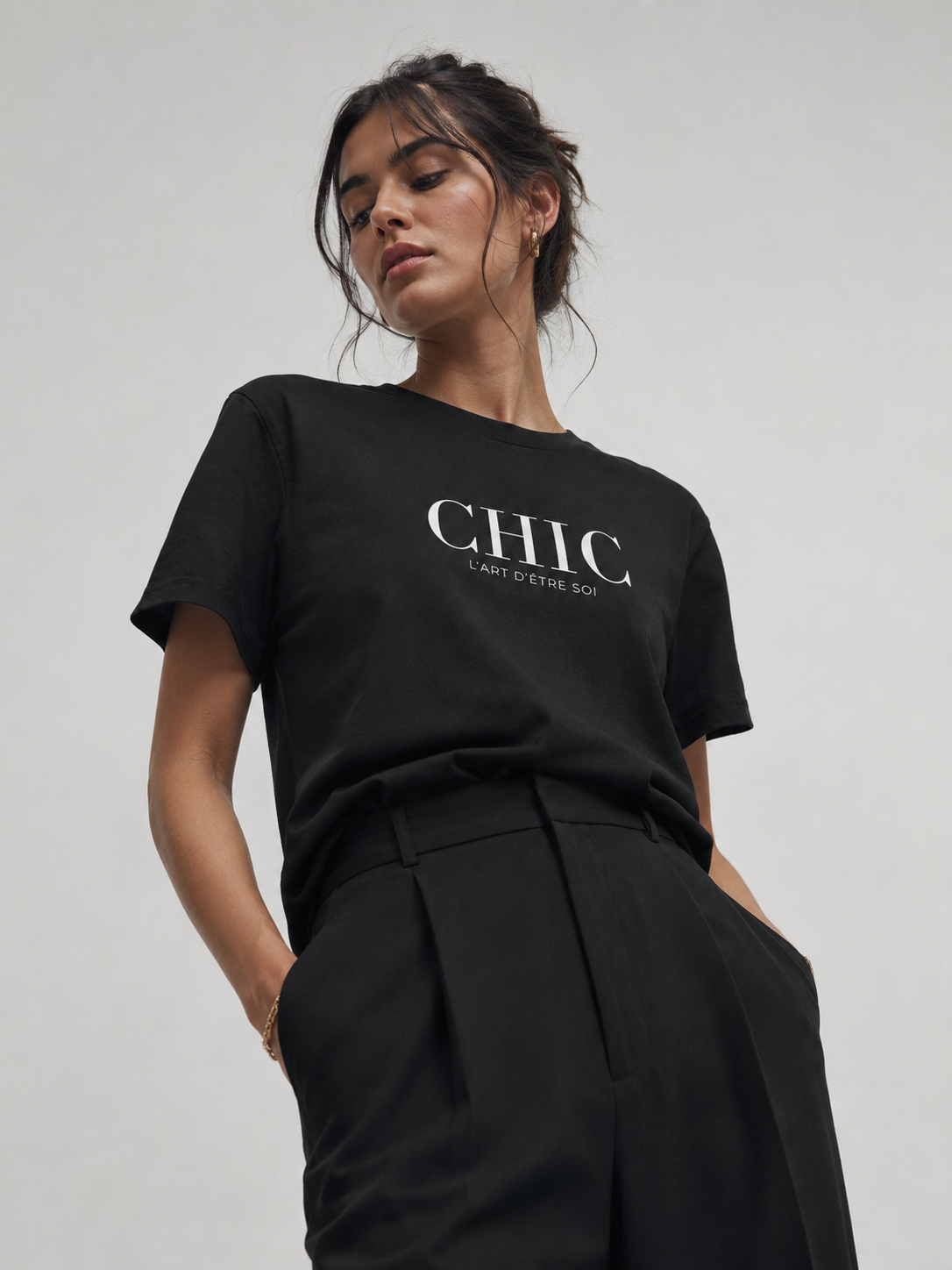 A woman wearing a black short-sleeve t-shirt with 'CHIC L'art D'etre soi' printed in white across the chest, paired with black pleated trousers, against a plain light gray background.